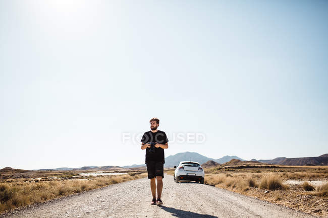Бородатый мужчина держит камеру и идет по пустынной дороге от припаркованной машины
. — стоковое фото