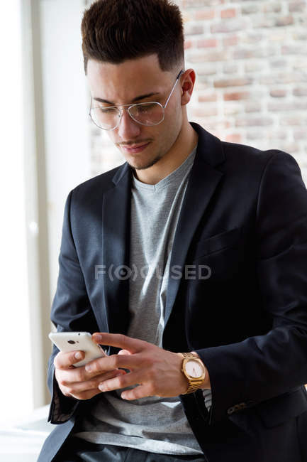 Porträt eines jungen Geschäftsmannes, der Smartphone an Ziegelwand benutzt. — Stockfoto