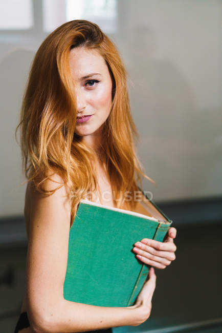 Portrait of redhead topless woman covering breasts with book and looking at camera. — Stock Photo