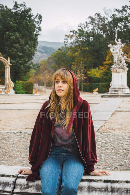 Brunette fille en sweat à capuche rouge assis sur parapet sur des statues sur fond — Photo de stock
