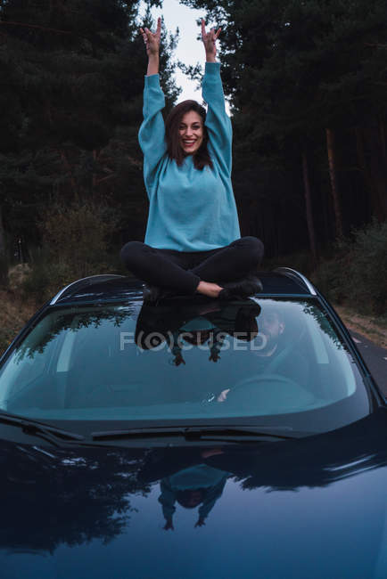 Chica en sudadera azul sentada en la parte superior del coche con las piernas cruzadas y los brazos levantados - foto de stock