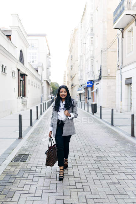 Улыбающаяся элегантная деловая женщина с сумочкой, гуляющая по улице и смотрящая в камеру
. — стоковое фото