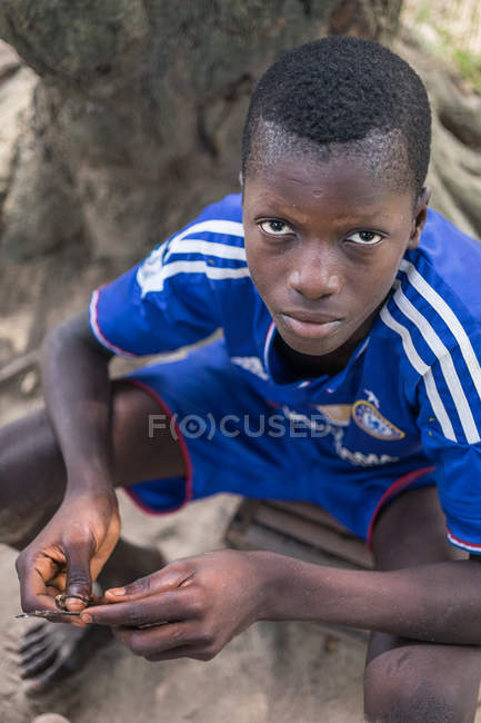 BENIN, AFRICA - AUGUST 31, 2017: Portrait of thoughtful boy sitting by tree and looking at camera — Stock Photo