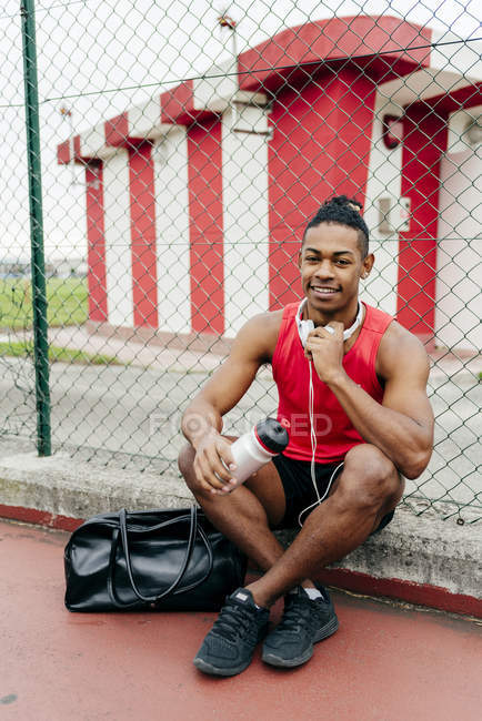 Esportista com fones de ouvido no pescoço sentado no playground com garrafa após o treino — Fotografia de Stock