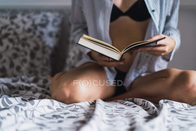 Chica de las cosechas en lencería y camisa sentado en la cama y sosteniendo el libro en las manos - foto de stock