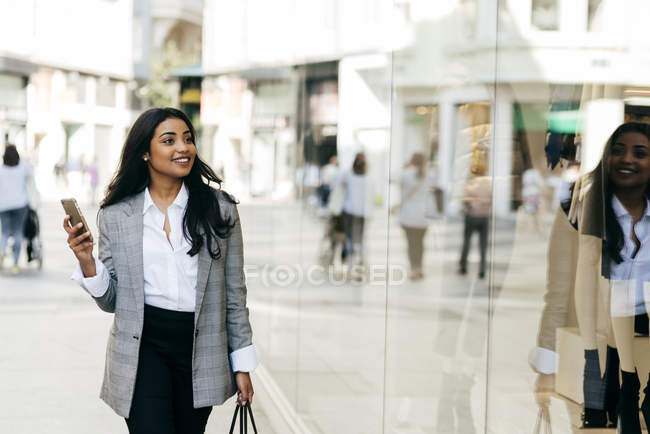 Retrato de mujer elegante con chaqueta caminando con smartphone en la mano a lo largo de los escaparates - foto de stock