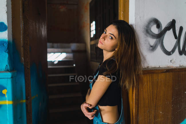 Vista lateral da menina morena posando no prédio abandonado e olhando para a câmera — Fotografia de Stock