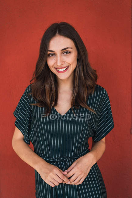 Portrait de fille brune souriante en robe rayée posant sur le mur rouge et regardant la caméra — Photo de stock