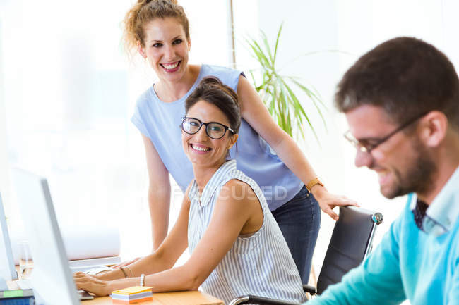 Dos mujeres sonrientes mirando a su compañero de trabajo en la oficina moderna . - foto de stock