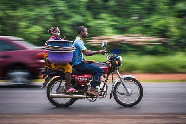 BENIN, ÁFRICA - 31 de agosto de 2017: Vista lateral de una mujer negra sentada detrás de un hombre que conduce una motocicleta en la carretera y mira la cámara - foto de stock