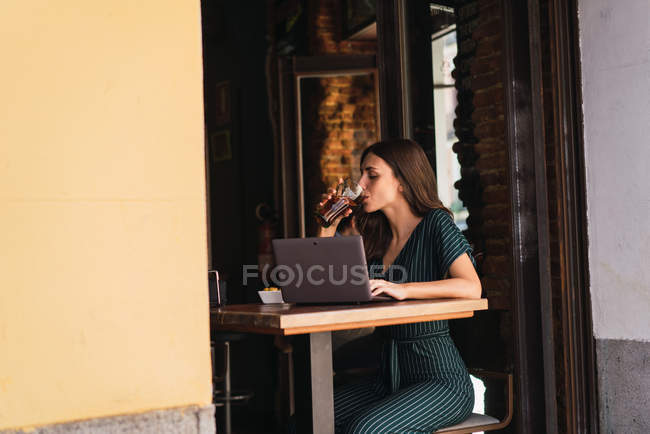 Vue latérale de la femme buvant de la coke à un ordinateur portable sur la table dans un café — Photo de stock
