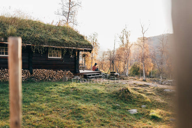 Женщина, сидящая на террасе рядом с деревянным домом в лесу — стоковое фото