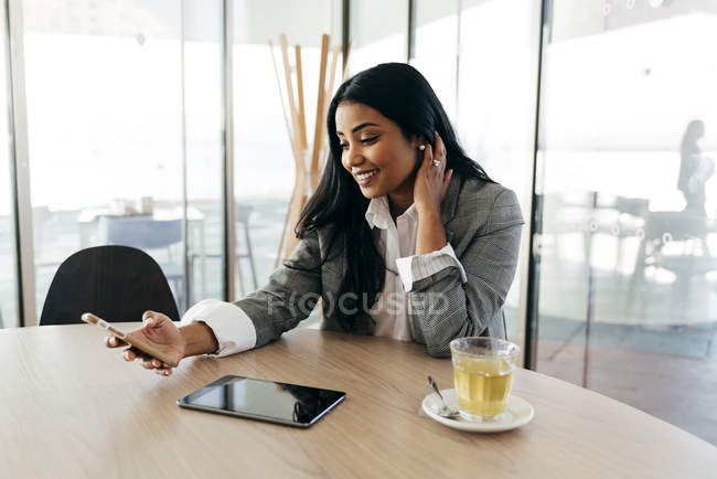 Портрет улыбающейся деловой женщины, сидящей за столом с чашкой чая и просматривающей смартфон — стоковое фото