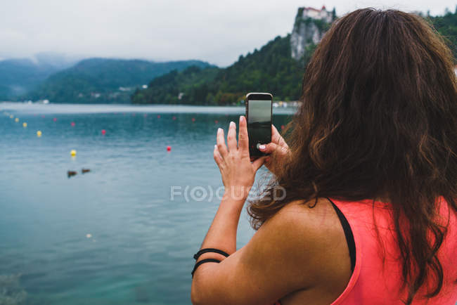 Visão traseira de mulher tomando tiros com smartphone de lago em montanhas . — Fotografia de Stock