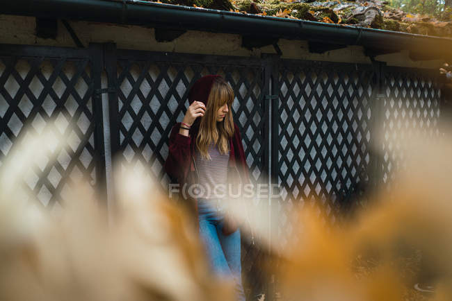 Brunette girl in hood romantically leaning on fence in park and adjusting hair — Stock Photo