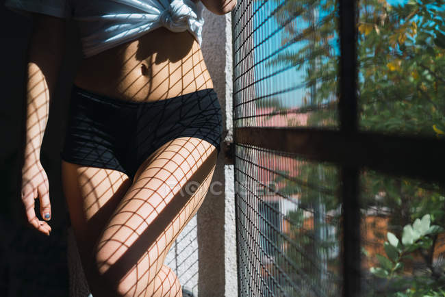 Crop attractive woman wearing black panties standing and posing at grid window. — Stock Photo