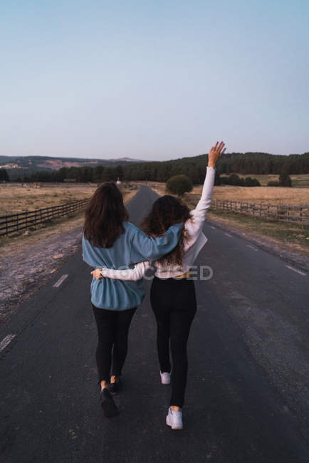 Rückansicht von zwei Frauen, die sich umarmen und ländliche Straße entlanggehen. — Stockfoto