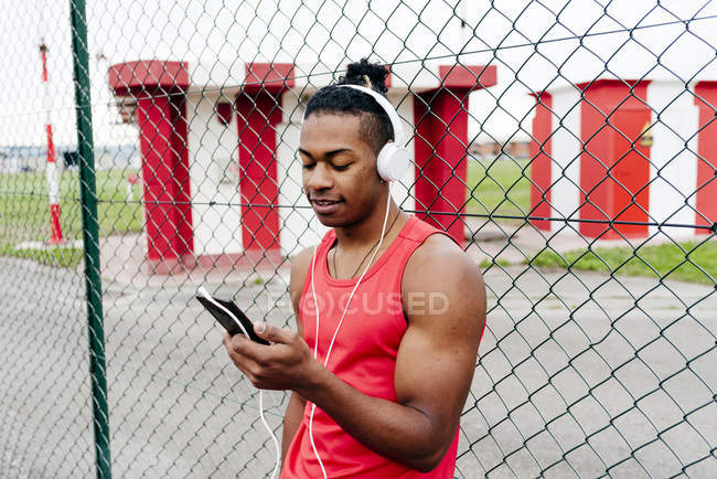Portrait of sportsman in headphones leaning on fence and browsing smartphone — Stock Photo
