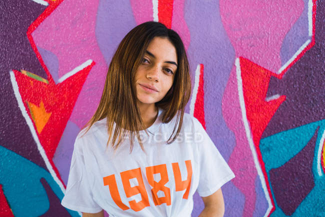 Brunette woman looking at camera over wall with graffiti on backdrop — Stock Photo