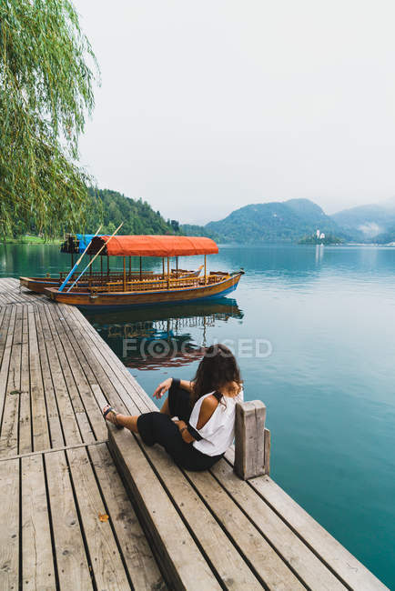 Задний вид женщины, сидящей на пирсе и смотрящей на холмы над озером — стоковое фото