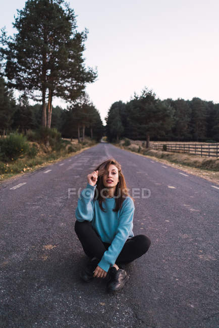 Retrato de menina em camisola azul sentado na estrada e tocando o cabelo — Fotografia de Stock
