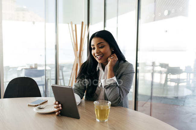 Smiling businesswoman sitting at table with cup of tea and looking at tablet in hand — Stock Photo