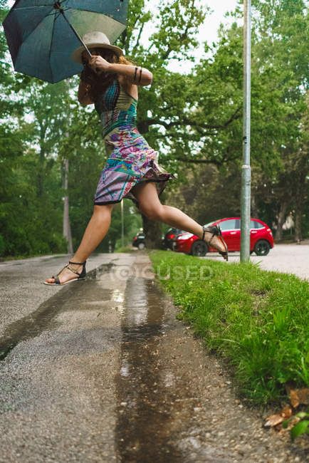 Vista laterale della donna con ombrello che salta sopra la pagaia sul marciapiede — Foto stock