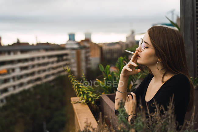 Vista lateral de mujer morena en gafas fumando cigarrillo en la terraza - foto de stock