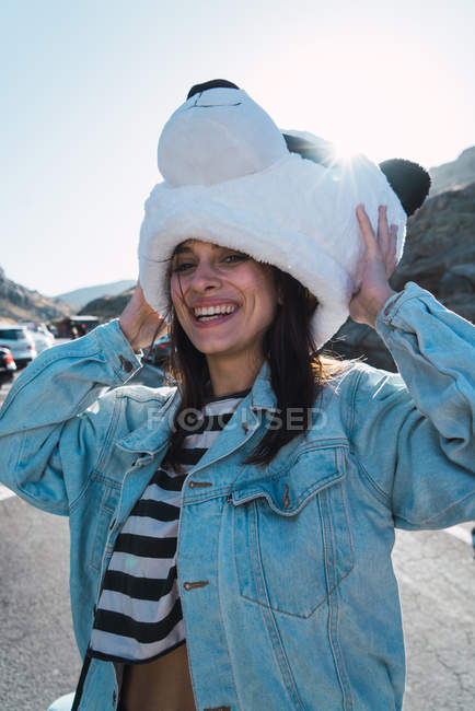 Fille riante en manteau de denim debout sur la route et portant la tête de jouet panda — Photo de stock
