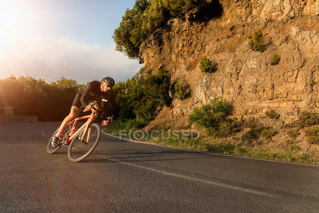 Cyclist riding bicycle along asphalt road near clay cliff on sunny day. — Stock Photo