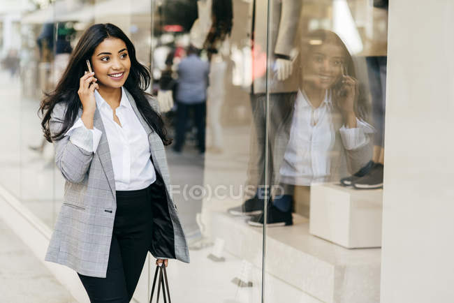 Mujer de negocios sonriente hablando en el teléfono inteligente y caminando por los escaparates - foto de stock