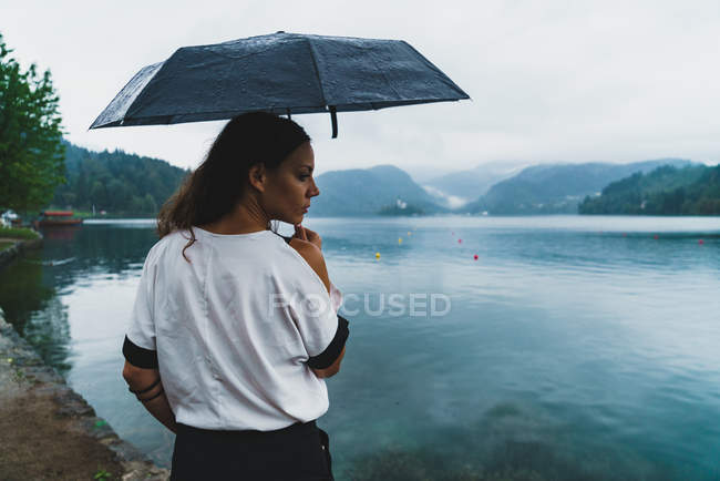 Mujer de pie en la orilla del lago con paraguas y mirando por encima del hombro - foto de stock