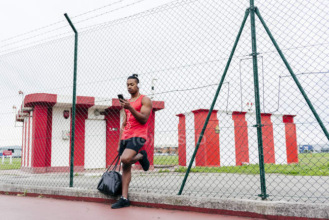 Sportler mit Kopfhörern lehnt sich an Drahtzaun und surft Smartphone — Stockfoto
