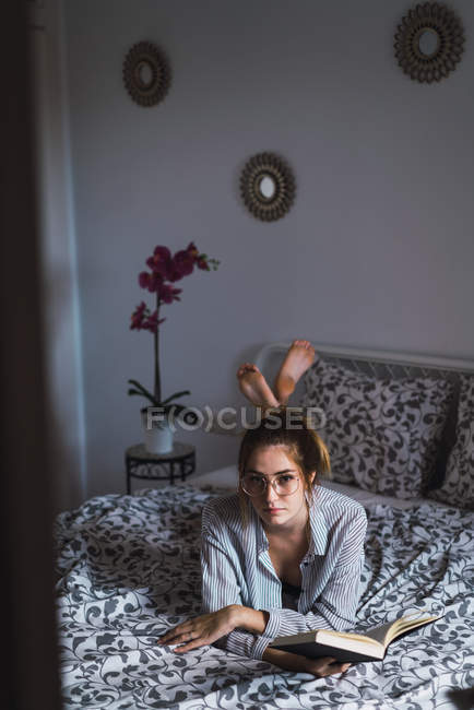 Attractive woman in eyeglasses lying on bed with book and looking at camera — Stock Photo