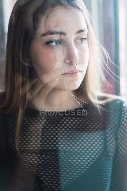 Ritratto di giovane ragazza con piercing posa pensierosa dietro vetro e distogliendo lo sguardo tristemente — Foto stock