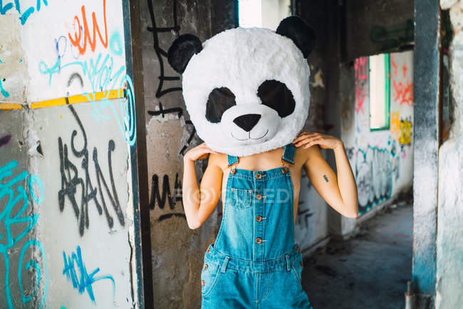 Portrait of girl in overall  with plush panda head posing at abandoned building — Stock Photo
