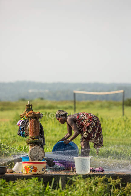BENIN, AFRIQUE - 31 AOÛT 2017 : Vue de côté d'une femme noire ethnique lavant des ustensiles au point d'eau sur fond de tropiques . — Photo de stock