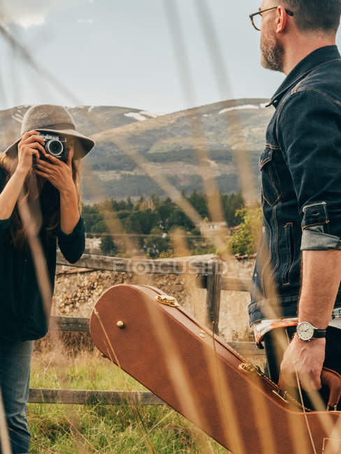 Woman with vintage camera taking shot of man with guitar case at countryside field — Stock Photo