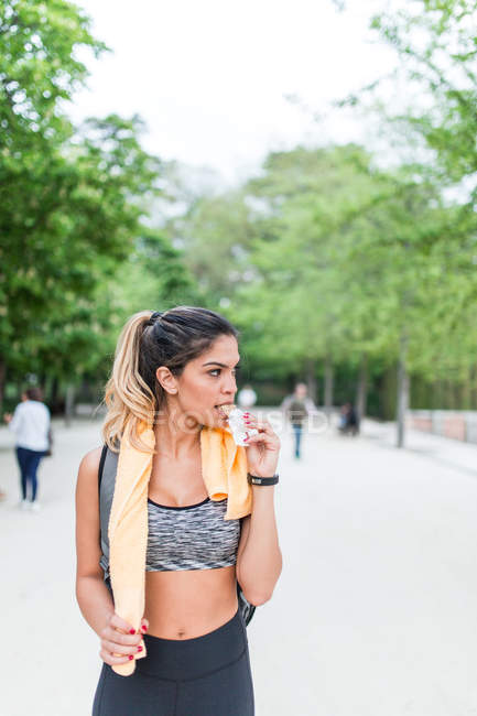 Retrato de chica en forma caminando por el callejón del parque después del entrenamiento y comiendo bocadillos - foto de stock