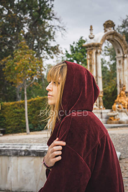 Side view of brunette girl in red hood posing over arch statue on background — Stock Photo