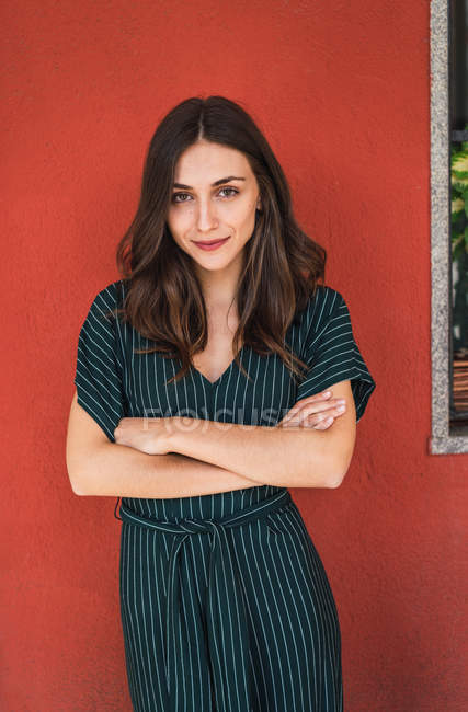 Portrait of brunette girl posing with arms crossed over red wall and looking at camera — Stock Photo