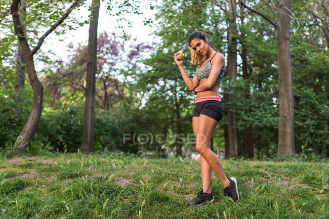 Athletic girl wearing sport outfit posing with apple in hand on green park lawn — Stock Photo
