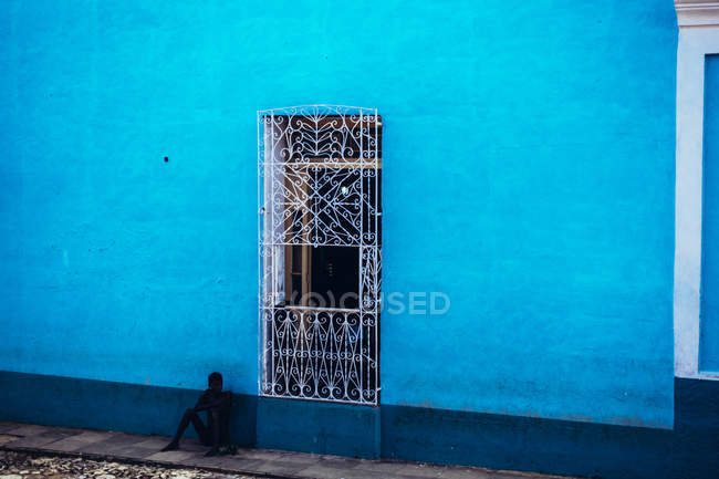 CUBA - 27 AOÛT 2016 : Homme assis sur le trottoir appuyé sur un mur bleu avec porte en métal ornemental . — Photo de stock