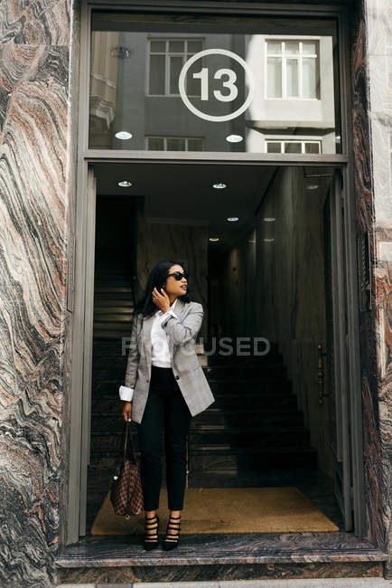 Portrait of elegant businesswoman in sun glasses walking out of building and looking aside — Stock Photo