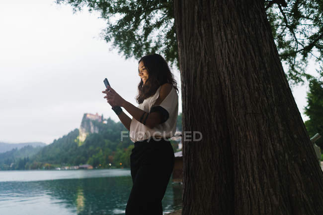 Vista lateral da menina morena em pé ao lado da árvore e smartphone de navegação — Fotografia de Stock