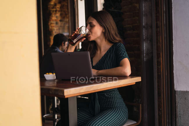 Side view of woman drinking coke at cafe table with laptop — Stock Photo