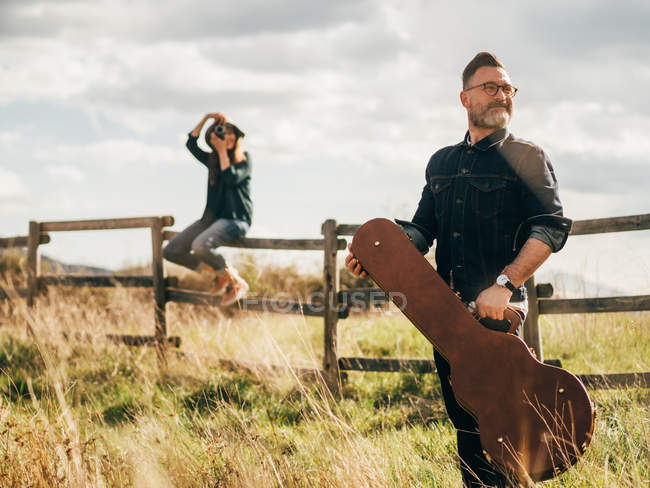 Frau mit Oldtimer-Kamera sitzt auf ländlichem Zaun und fotografiert Mann, der mit Gitarrenkoffer posiert und wegschaut — Stockfoto