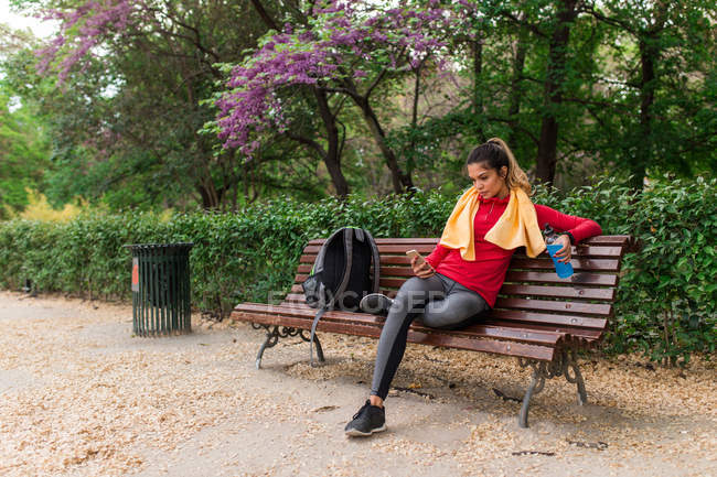 Спортивная девушка с бутылкой воды и полотенцем на плечах сидит в парке и просматривает смартфон — стоковое фото