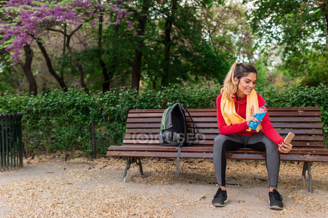 Sportive girl sitting on park bench with energetic drink and taking selfie — Stock Photo