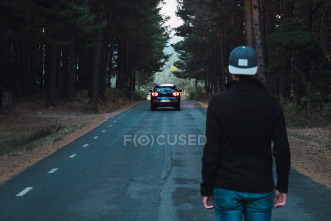 Parte trasera del hombre en la tapa de pie en la carretera forestal y mirando coche de conducción . - foto de stock